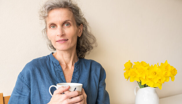 Middle aged woman with grey wavy hair and blue top seated with cup next to daffodils, looking at camera with half smile (selective focus)
