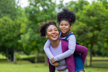 African American mother is playing piggyback riding with her young daughter while having a summer picnic in the public park for wellbeing and happiness concept