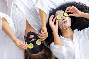 Couple of girlfriend in bathrobe doing skincare routine using facial mask and cucumber slice on spa...
