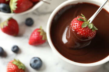 Dipping strawberry into fondue pot with chocolate on white table, top view