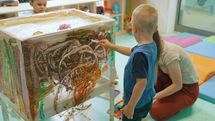 Cling film painting. Toddler painting with a sponge, brushes and paints on a cling film wrapped all the way round the wooden shelf unit. A teacher helping them. Creative activity for kids sensory