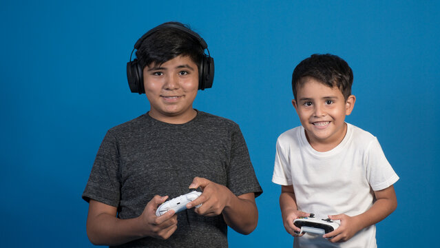 Retrato de estudio de dos niños jugando a videojuegos con auriculares y joystick. Posando en fondo Azul.