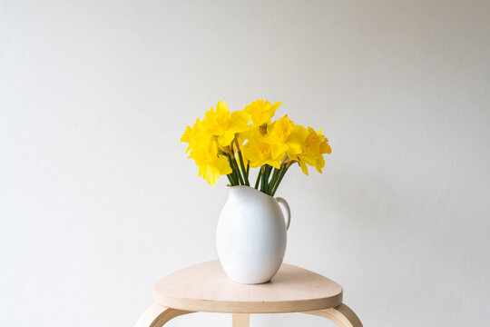 Closeup of daffodils in white vase on small wooden table against beige wall (selective focus)