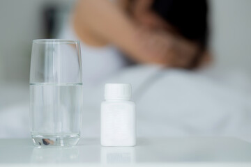 Young asian woman is depression and having mental health problem sitting on bed while drug and water on desk in the bedroom, emotion and negative, woman and expression, medical and health concept.