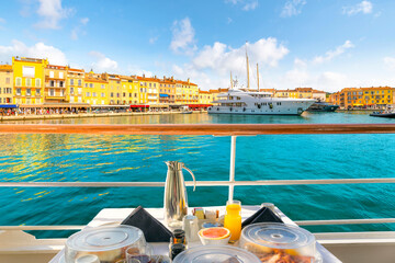 View of the colorful old town and marina of the French Riviera resort town of Saint-Tropez, France, from a deck balcony with full breakfast setup on a luxury yacht. - obrazy, fototapety, plakaty