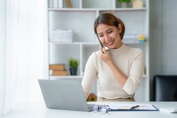 Fototapeta premium Young Asian businesswoman sit with her laptop computer happily talking on the phone with her customer explaining the detail in the office.