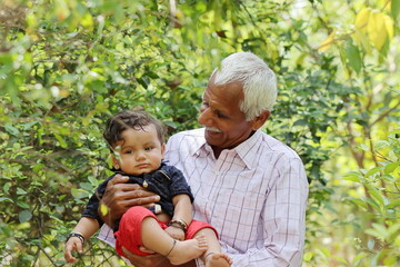 Portrait photo of Indian Senior grandfather with toddler grandson standing in green garden with blur background