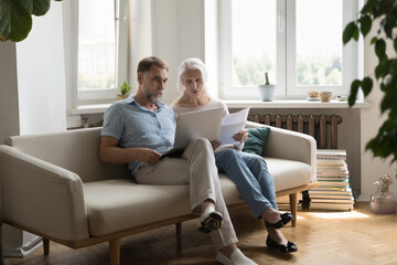 Serious older 60s couple paying paper bills on Internet, using online banking app on laptop, checking documents together, counting loan, insurance, tax fees, planning budget, discussing expenses