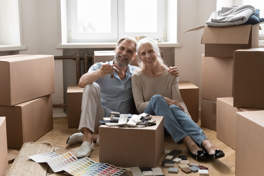 Senior 60s married couple enjoying moving into new apartment, sitting on floor among cardboard boxes, renovation samples, holding keys, hugging, looking at camera, smiling. Home portrait
