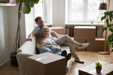 Older senior couple of new landlords, homeowners resting on sofa in room with cardboard packaging...