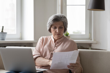 Focused senior elder accountant woman checking paper invoice at laptop, paying bills, loan, insurance fees, counting budget, expenses, reading document, holding receipt, sitting on home sofa