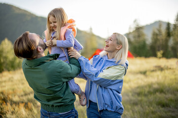 Young caucasian couple with little girl and dog have fun while hiking in the mountains. Happy family spend summer vacation at campsite. Father tossing daughter in his arms © rh2010