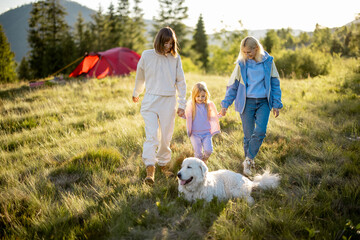 Two women walk with little girl and white dog, happily spending summer time on nature. Homosexual family with a kid travel in mountains
