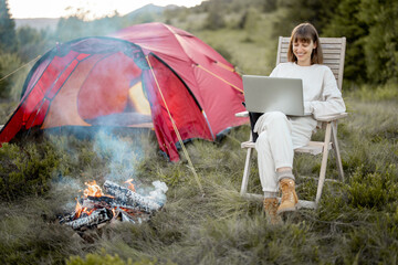 Young woman works on laptop while sitting relaxed on chair by the campfire, traveling with tent in...