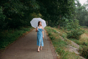 Fototapeta na wymiar A slender young girl with blond long hair, in a blue summer dress and shoes, walking with a white umbrella in her hands, on a path in a forest park area in the rain.