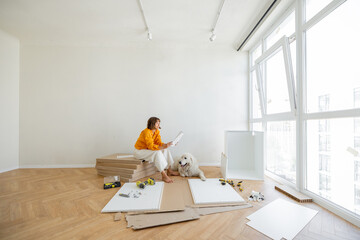 Young woman assembles furniture by herself, sitting with dog near window and reading manual at new...