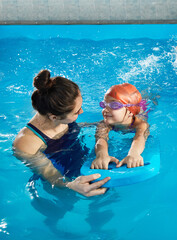 Little girl learning to swim in indoor pool with pool board and trainer