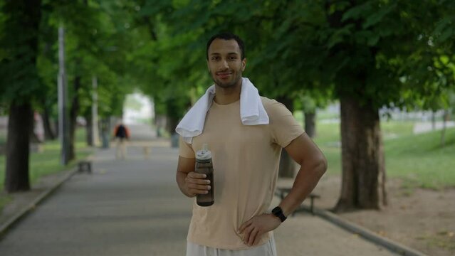 Portrait of the afro american man looking at the camera holding sport bottle wearing smart watch carries a towel around his neck in the park after the training. Sport and healthy lifestyle concept.