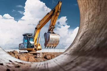 Fotobehang A powerful caterpillar excavator digs the ground against the blue sky. Earthworks with heavy equipment at the construction site. View from a large concrete pipe. © Anoo