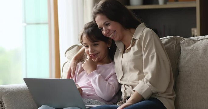 Cheerful mom and sweet little daughter kid watching, commenting, discussing cartoon movie on laptop, using computer for video call, speking, talking, smiling, laughing