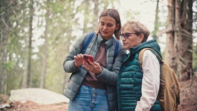 Portrait of a senior caucasian woman and her adult daughter looking at the map on the smartphone while hiking in the woods together.