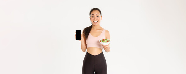 Sport, wellbeing and active lifestyle concept. Smiling slim and cute asian fitness girl, gym coach showing salad and smartphone screen, recommend download diet tracker or calories reminder