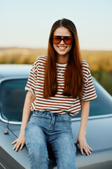A fashionable woman smiles sweetly in stylish sunglasses, a striped t-shirt and jeans, sits on the trunk of a car and looks at the beautiful nature of autumn. Travel lifestyle