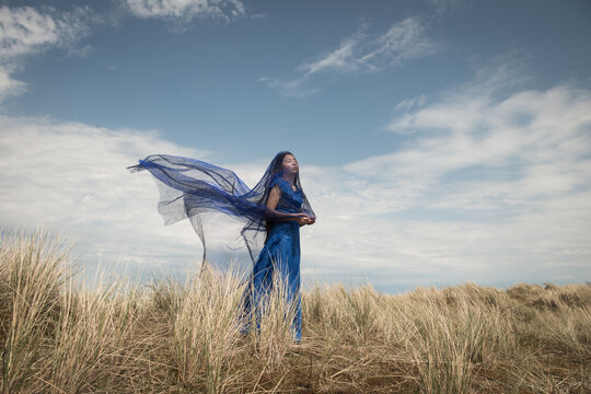 fine art portrait of woman in blue dress standing in dunes  coverd by thin fabric