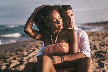 Multiethnic couple of young lovers relaxing together hugging on the beach by the ocean - travel, tourism and romantic people in tropical destination lifestyle concept