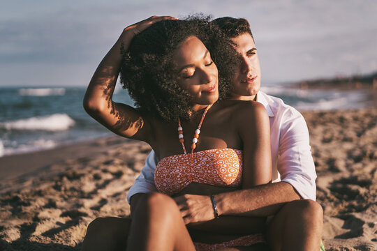 Multiethnic couple of young lovers relaxing together hugging on the beach by the ocean - travel, tourism and romantic people in tropical destination lifestyle concept - Powered by Adobe