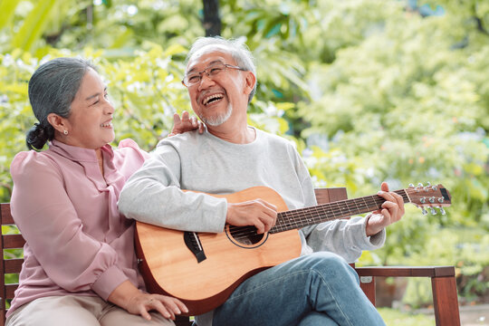 Happy Asian senior couple elderly man playing guitar music song enjoy having fun happiness outside house near backyard garden. Retirement insurance concept