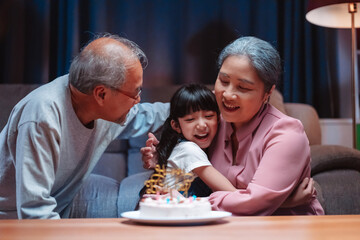 Asian happy family of little girl hugging lover with grandparents after blowing out candles on cake. Celebrate birthday anniversary party on table at night in living room, having happiness lifestyle.