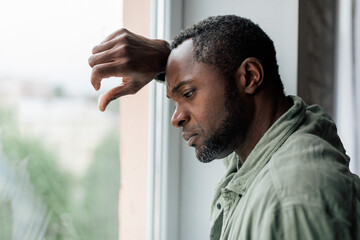 Unhappy frustrated adult african american guy in casual suffering from depression and bad news near window - obrazy, fototapety, plakaty