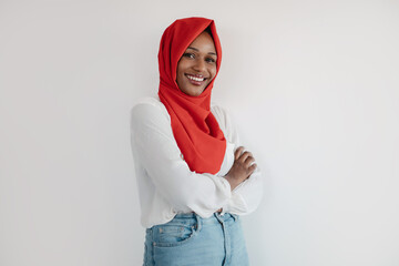 Portrait of black woman in hijab posing with folded arms on light studio background and smiling at...