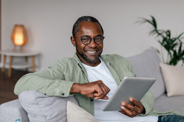 Smiling mature african american male in glasses and casual watching video on tablet, chatting in...