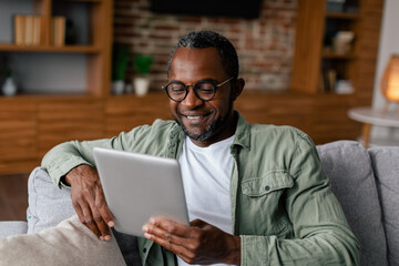 Cheerful adult african american man in glasses and casual watching video on tablet, chatting in living room interior