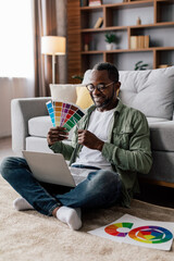 Fototapeta premium Cheerful adult black guy designer in glasses and casual show color palettes in laptop on floor in living room