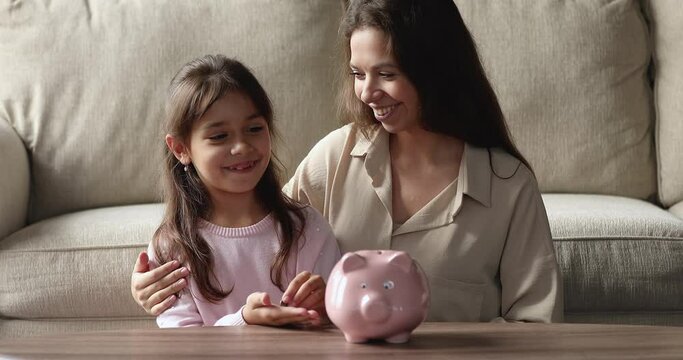 Happy young mom teaching little daughter kid to make reserve fund, saving money, inserting coins, cash into ceramic piggy bank, smiling, laughing. Financial education concept