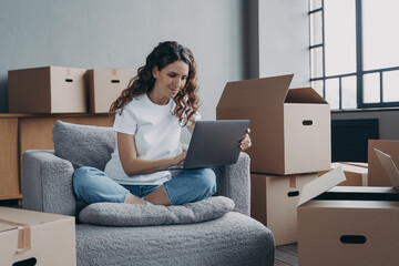 Female renter homeowner sitting on armchair with laptop orders removal service on moving day