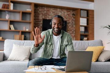 Glad mature african american male in casual and headphones waving hand, have video call on laptop