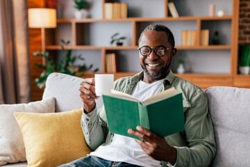 Cheerful adult african american man in glasses and casual reading book and enjoy favorite drink on...