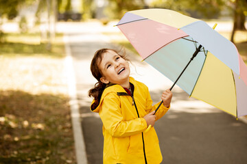 Little cute girl kid child in a yellow raincoat with multicolored rainbow umbrella laughs and looks...
