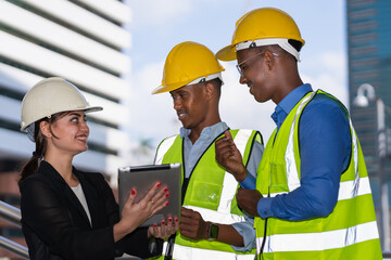 caucasian businesswoman in black suit wear helment working on tablet with black engineer colleague. group of engineer working at construction site outside building in city