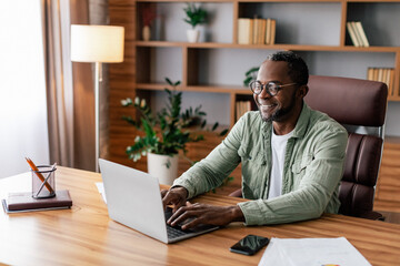 Cheerful confident mature african american businessman in glasses and casual working on laptop in...