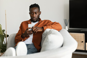 African american man sitting on couch, chatting with girlfriend or making online order via mobile app, using cellphone