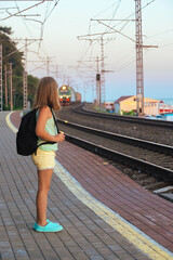 girl schoolgirl 8-10 years old travels by rail, she stands with a backpack on her shoulders and waits for a train on the platform of the station