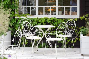 Victorian style white empty metal chairs and tables in outdoor cafe. Wrought iron furniture, old fashion cozy cafe terrace, restaurant tables waiting for customers at an outdoor terrace. Sidewalk cafe