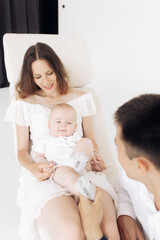 Young happy parents sit in armchair and play with their baby boy.