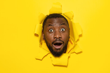 Shocked african american man reacting with surprisement, looking at camera, posing in torn yellow paper hole
