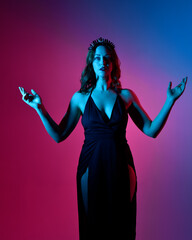 Close up portrait of beautiful woman model wearing elegant black dress and crown, posing against a studio background with fantasy inspired arm gestures, multi coloured creative lighting.